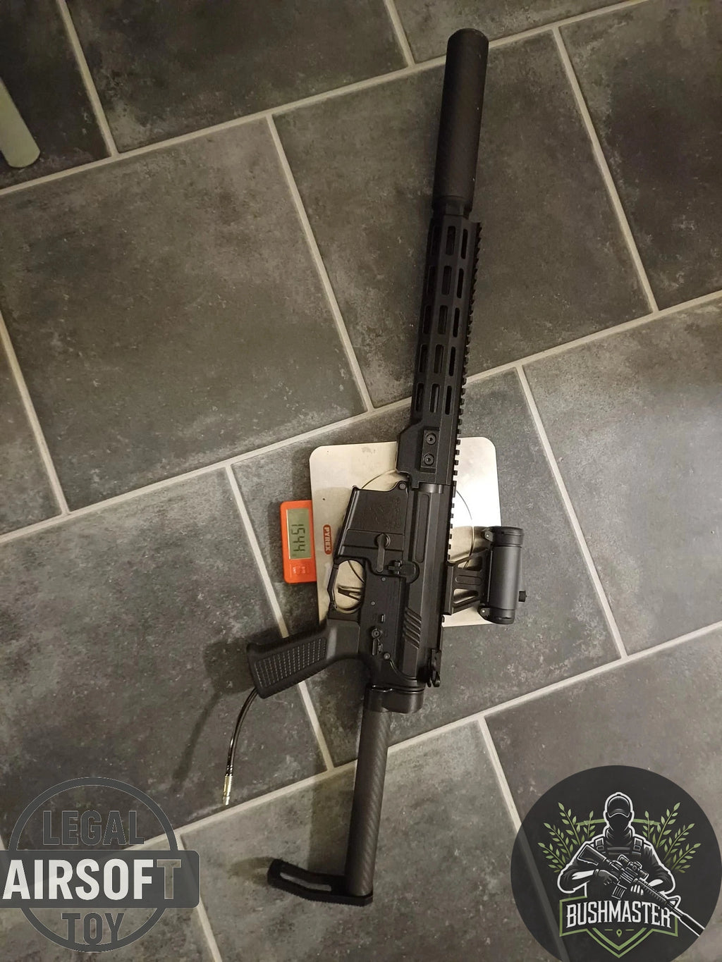 Black airsoft Bushmaster rifle with suppressor on tiled floor, placed on digital scale reading 1544 grams