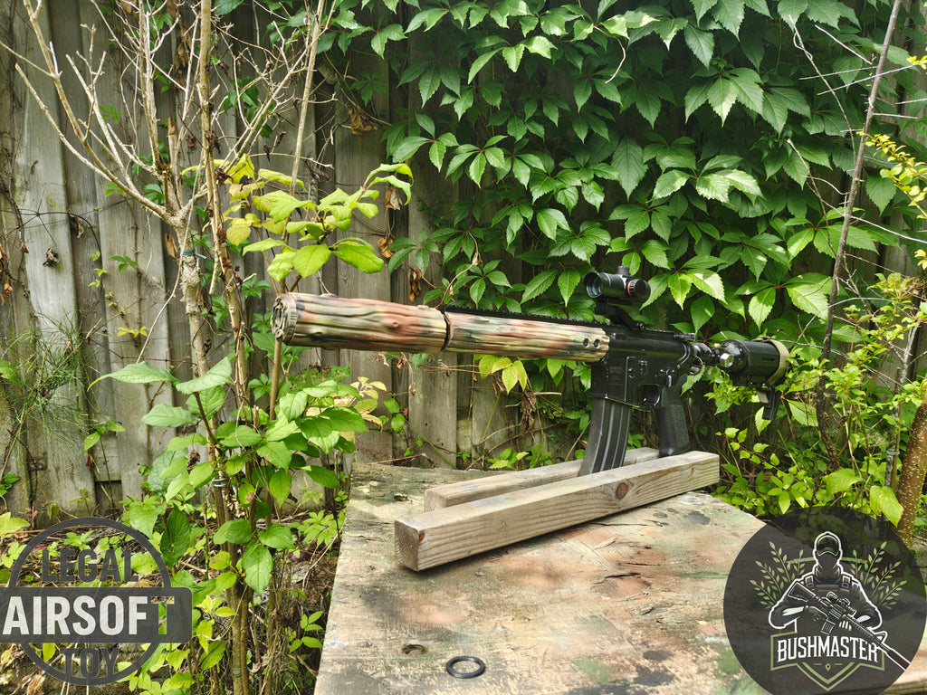 Camouflaged airsoft rifle with scope on weathered wooden table outdoors near leafy green plants