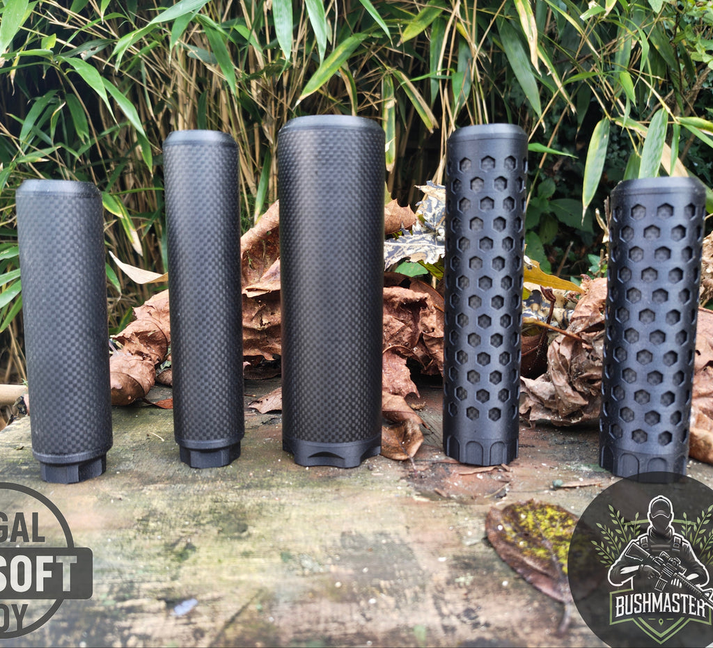 Five black airsoft suppressors with textured and honeycomb designs standing on a wooden surface outdoors with leaves and bamboo in background