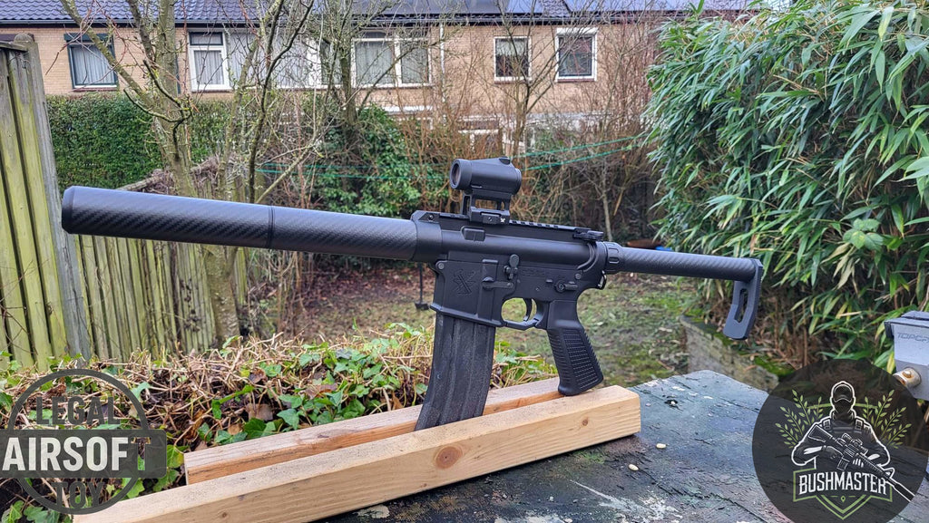 Black Bushmaster airsoft rifle with suppressor and red dot sight on wooden stand in outdoor garden setting